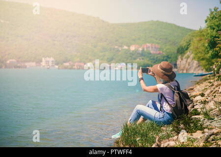 girl traveler sits on a mountain lake and takes pictures Girl takes photo for travel blog. View from back of the tourist traveler. Stock Photo