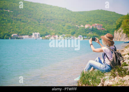 girl traveler sits on a mountain lake and takes pictures Girl takes photo for travel blog. View from back of the tourist traveler. Stock Photo