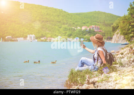 girl traveler sits on a mountain lake and takes pictures Girl takes photo for travel blog. View from back of the tourist traveler Stock Photo