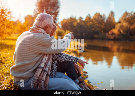 Senior couple having picnic by autumn lake. Happy man and woman enjoying nature and hugging. Man pointing into distance Stock Photo