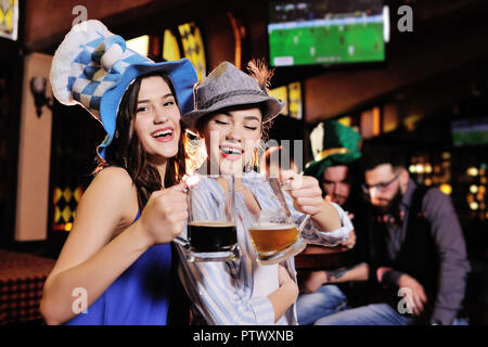 two young beautiful  girlfriends in Bavarian hats on the background of a bar holding a beer watching football on a TV monitor during the celebration o Stock Photo