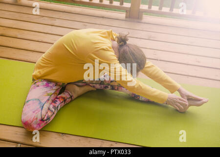 Mature woman, doing stretching exercises on her yoga mat, outside on the wood deck. Stock Photo