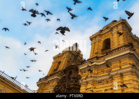 A flock of pigeons fly in circles against a blue sky around San Francisco Church, in Lima, Peru, a twin bell-tower cathedral that is painted yellow. Stock Photo