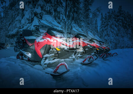 Polar night in Lapland, Finland. Red snowmobiles are ready for adventure ride. Vehicles parked in line near beautiful forest. Heavy snow. Winter seaso Stock Photo