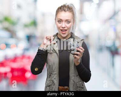 Young blonde woman taking pictures using vintage camera over isolated background with surprise face pointing finger to himself Stock Photo