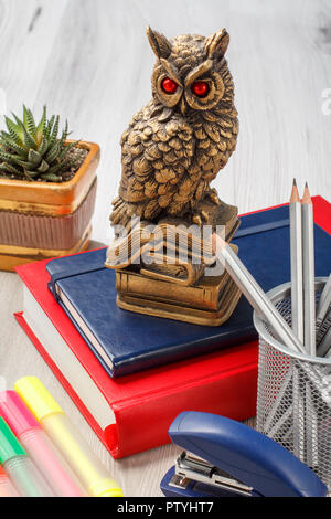 Books with statuette of owl on the top, metal stand with pencils, stapler, colored markers and houseplant in a pot on the background. School and offic Stock Photo