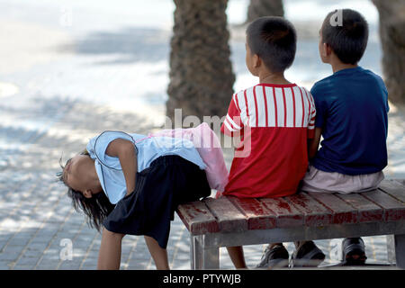 Funny kids. Three young friends sitting on a bench, with the young girl contorting to view something above. Thailand,  Southeast Asia Stock Photo