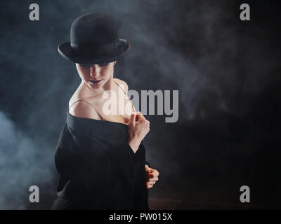 Young beautiful ballerina on smoke stage dancing modern ballet. performs smooth movements with hands against spotlights background. Woman in black cos Stock Photo