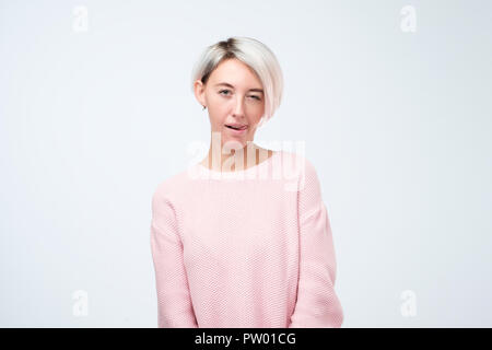 Cheerful young beautiful girl in pink sweater smiling winking showing tongue looking at camera over white background. Stock Photo