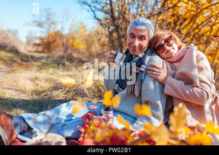 Senior couple having tea in autumn forest. Happy man and woman enjoying picnic and nature. Smiling man pouring tea out of thermos Stock Photo
