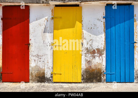 Red and yellow and blue old wooden doors on the shabby light wall background outdoors. Sun is shining on them. Horizontal. Stock Photo
