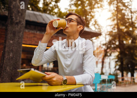 Young businessman enjoys coffee in outdoor cafe using tablet. Handsome guy drinking tea on city street holding tablet Stock Photo