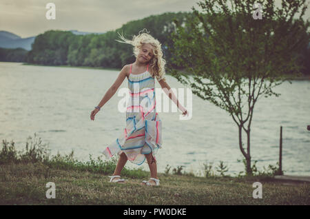 adorable little girl in dress at the pond outdoors in the afternoon having fun  at orange sunset back view Stock Photo
