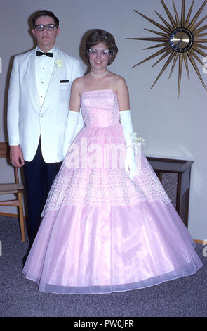 1962, America, going to the prom.....a young man wearing a white tuxedo and and a young woman wearing a long frilly pink dress stand together for a picture before they attend the prom, a traditional and important end of term dance and social event for US high school students. In the 50s and 60s, the prom was more of a couple's event, where a boy would ask a girl to be his date and would escort her after picking her up at her home and having lots of picture taken! Stock Photo