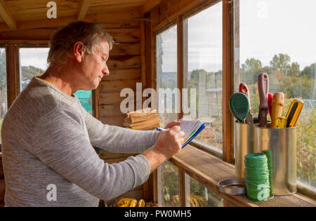 white male writing notes in garden shed, looking out onto allotments, small garden implements and twine on window ledge Stock Photo