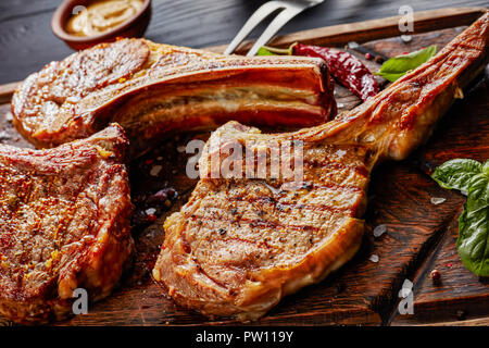 close-up of grilled steaks bone-in or cowboy beef steaks on an old rude wooden cutting board with fork on a black table with ketchup and mustard, view Stock Photo