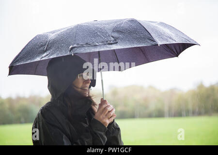 Kidderminster, UK. 12th October, 2018. UK weather: it's very wet and windy! Milder temperatures may be tempting us outdoors, but visitors to a Kidderminster country park have to brave heavy spells of rain and increasingly strong, gusty winds this morning. A young lady, in wet coat (hood up), is standing isolated in the pouring rain holding a wet open umbrella over her, watching the rain. Credit: Lee Hudson/Alamy Live News Stock Photo