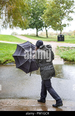 Kidderminster, UK. 12th October, 2018. UK weather: it's very wet and very windy! Milder temperatures may be tempting us outdoors, but visitors to a local park in Kidderminster have to brave heavy spells of rain and increasingly strong, gusting winds this morning. A lady, in wet rain coat, is isolated here struggling in the windy weather - her umbrella being blown inside out! Credit: Lee Hudson/Alamy Live News Stock Photo