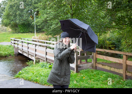 Kidderminster, UK. 12th October, 2018. UK weather: it's very wet and very windy! Milder temperatures may be tempting us outdoors, but visitors to a park in Kidderminster have to brave heavy spells of rain and increasingly strong, gusting winds this morning. A drenched lady, in wet rain coat, is isolated here struggling in the windy weather with her umbrella. Credit: Lee Hudson/Alamy Live News Stock Photo