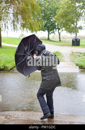 Kidderminster, UK. 12th October, 2018. UK weather: it's very wet and very windy! Milder temperatures may be tempting us outdoors, but visitors to a park in Kidderminster have to brave heavy spells of rain and increasingly strong, gusting winds this morning. A lady (from behind) in wet rain coat, is isolated here struggling in the windy weather - her umbrella being blown inside out! Credit: Lee Hudson/Alamy Live News Stock Photo