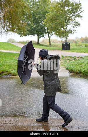 Kidderminster, UK. 12th October, 2018. UK weather: it's very wet and very windy! Milder temperatures may be tempting us outdoors, but visitors to a local park in Kidderminster have to brave heavy spells of rain and increasingly strong, gusting winds this morning. A drenched lady, in wet rain coat, is isolated here struggling in the windy weather with her umbrella. Credit: Lee Hudson/Alamy Live News Stock Photo