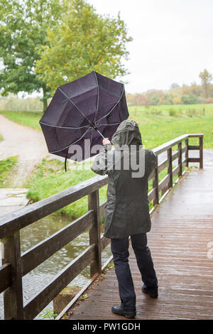 Kidderminster, UK. 12th October, 2018. UK weather: it's very wet and very windy! Milder temperatures may be tempting us outdoors, but visitors to a park in Kidderminster have to brave heavy spells of rain and increasingly strong, gusting winds this morning. A lady (from behind) in wet rain coat, is isolated here struggling in the windy weather - her umbrella being blown inside out! Credit: Lee Hudson/Alamy Live News Stock Photo