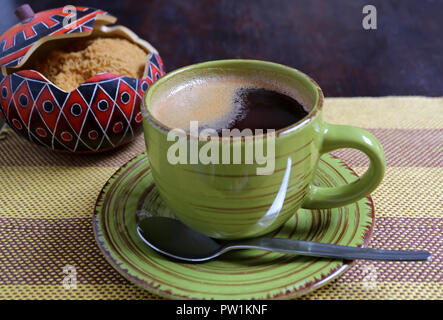 Hot coffee in a green cup with blurred sugar pot of brown sugar in backdrop served on stripe tablecloth Stock Photo