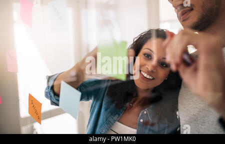 Business couple writing on sticky notes pasted on transparent glass wall in office. Office colleagues discussing business ideas and plans on a transpa Stock Photo