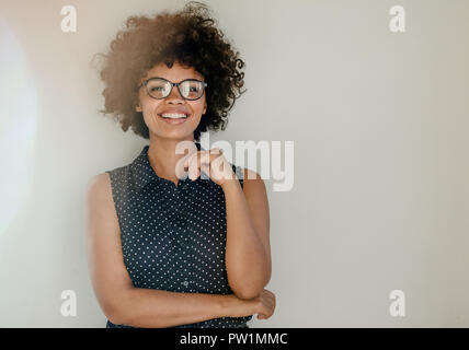 Portrait of beautiful young woman in casuals standing by a wall and smiling. Happy young african female with curly hair wearing spectacles looking at  Stock Photo