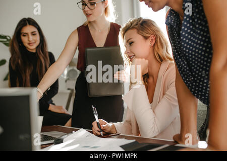 Group of young businesswomen in casuals looking at laptop and discussing work. Multi-ethnic female coworkers working together in office. Stock Photo