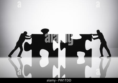 Silhouette Of Two Businesspeople Joining Jigsaw Puzzle Pieces On Reflective Desk Stock Photo