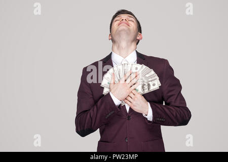 Dream come true! satisfied young businessman in classic violet coat standing, proud with closed eyes and lean on fan of money to the chest and smile.  Stock Photo
