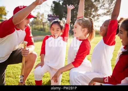 Girl baseball team kneeling with their coach, raising hands Stock Photo