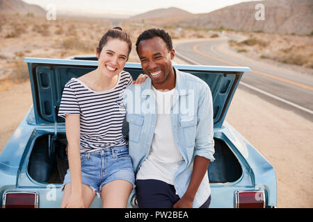 Portrait Of Couple Sitting In Trunk Of Classic Car On Road Trip Stock Photo