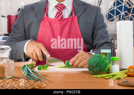 A businessman, father, husband in suit and red tie wearing red apron and cutting broccoli and vegetables with a knife on a wooden cutting board Stock Photo