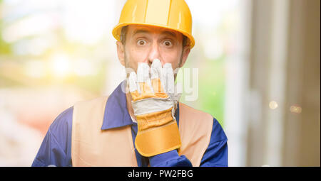Senior engineer man, construction worker covers mouth in shock, looks shy, expressing silence and mistake concepts, scared Stock Photo