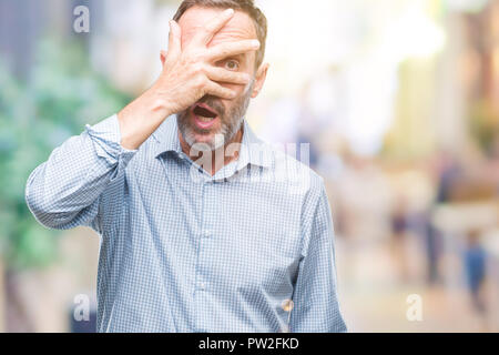 Middle age hoary senior business man over isolated background peeking in shock covering face and eyes with hand, looking through fingers with embarras Stock Photo