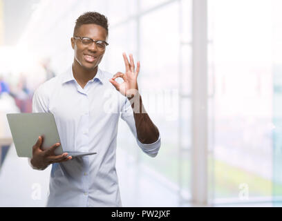 Young african american businessman using computer laptop doing ok sign with fingers, excellent symbol Stock Photo