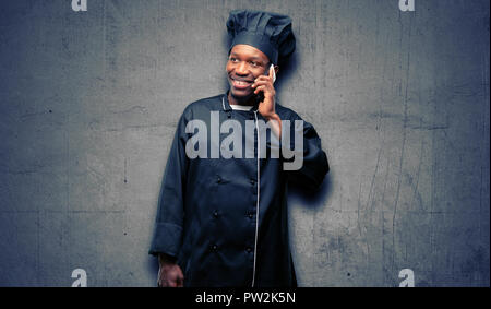 Young black cook wearing chef hat happy talking using a smartphone mobile phone Stock Photo