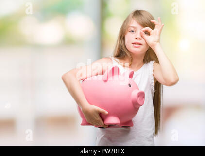 Young blonde toddler holding piggy bank with happy face smiling doing ok sign with hand on eye looking through fingers Stock Photo