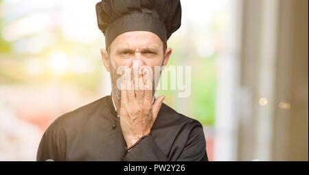 Senior cook man, wearing chef hat covers mouth in shock, looks shy, expressing silence and mistake concepts, scared Stock Photo