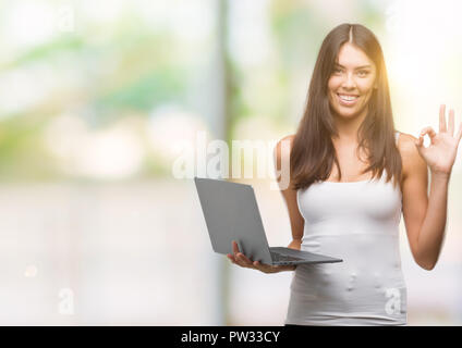 Young hispanic woman using computer laptop doing ok sign with fingers, excellent symbol Stock Photo