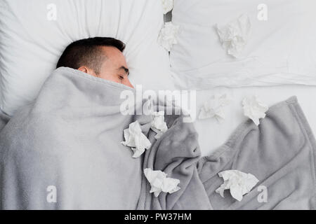 high angle view of an ill young caucasian man in bed, covered with a light gray blanket, surrounded by used tissues Stock Photo