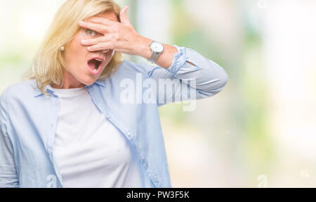 Middle age blonde woman over isolated background peeking in shock covering face and eyes with hand, looking through fingers with embarrassed expressio Stock Photo