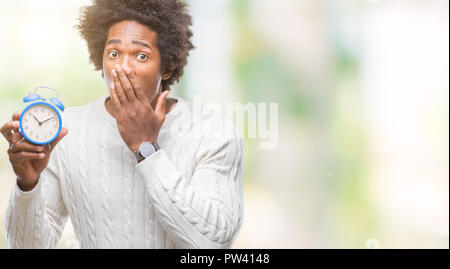Afro american man holding vintage alarm clock over isolated background cover mouth with hand shocked with shame for mistake, expression of fear, scare Stock Photo