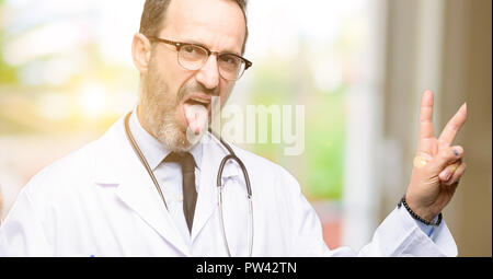 Doctor senior man, medical professional looking at camera showing tong and making victory sign with fingers Stock Photo