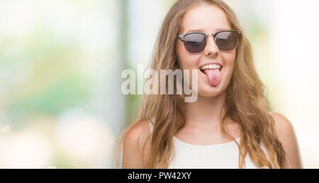 Young blonde woman wearing sunglasses sticking tongue out happy with funny expression. Emotion concept. Stock Photo