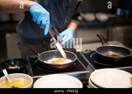 Hotel chef hands with gloves cooking fried eggs on hot pan for breakfast in restaurant at hotel. Stock Photo