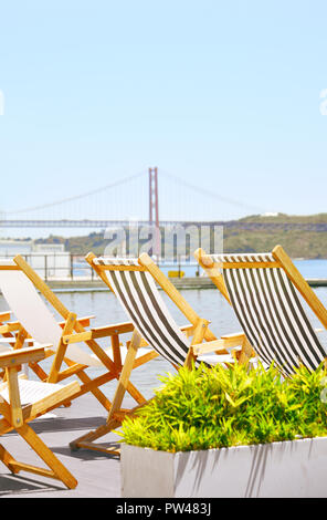 Three chaise long looking out to the sea and the 25 Abril bridge in Lisbon, Portugal on a bright summer day Stock Photo