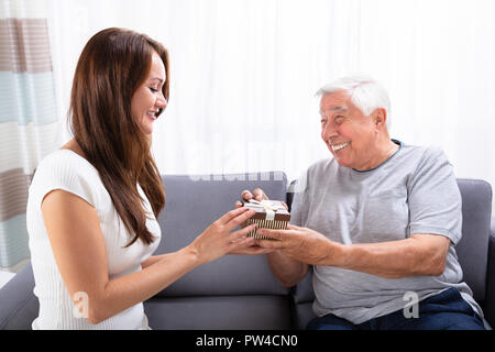Woman Giving Gift To Her Happy Father Sitting On Sofa Stock Photo